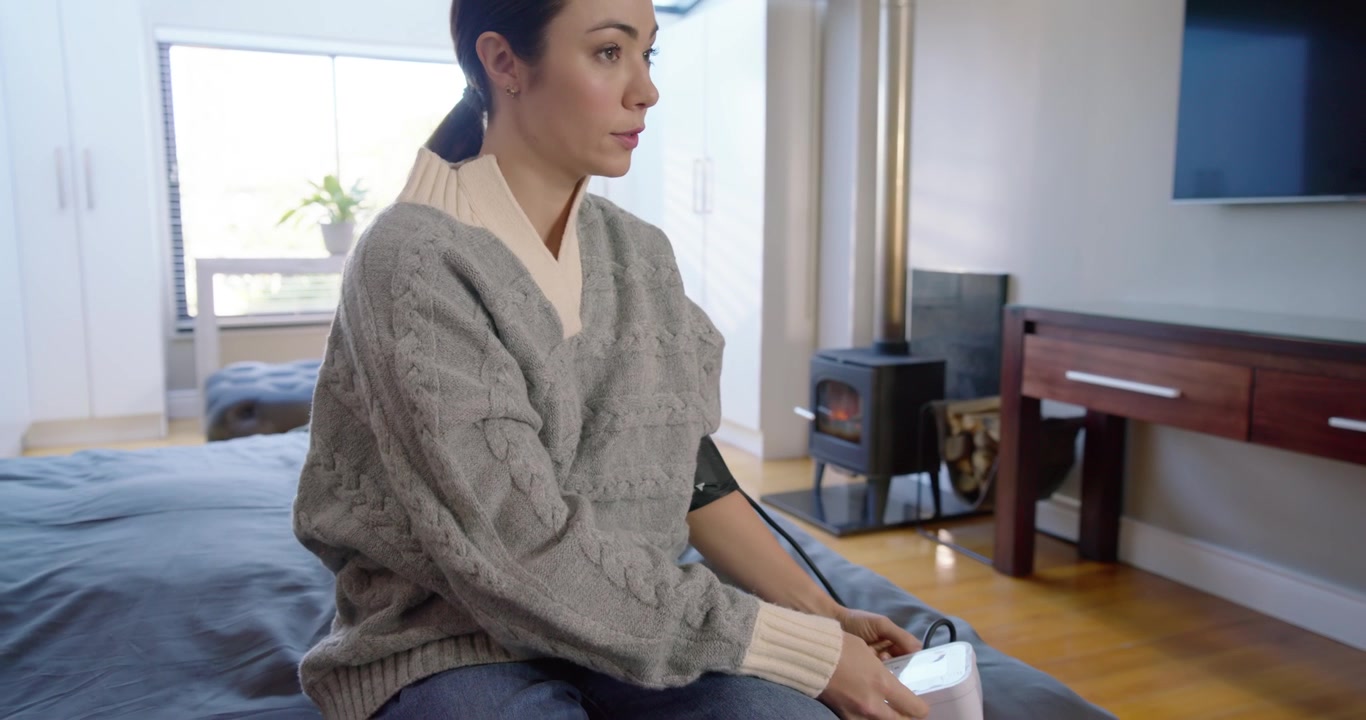 Donna con un maglione grigio seduta sul letto che preme il monitor, regola il bracciale e controlla la pressione sanguigna