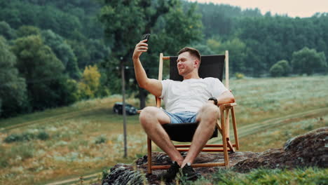 freelancing man working in the nature having video chat on his phone. man holds his phone up and talks sitting in a chair on the rock.