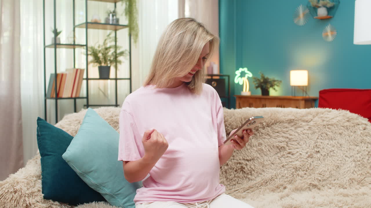 Pregnant woman celebrating baby news texting on smartphone making victory sign smiling super indoors