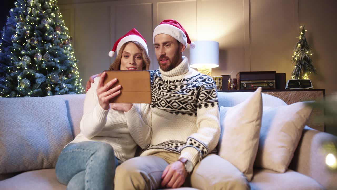feliz pareja joven casada hablando en una videollamada en línea en una tableta mientras se sienta en una habitación acogedora con un árbol resplandeciente decorado con navidad