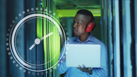 animation of clock over african american man working with laptop and servers