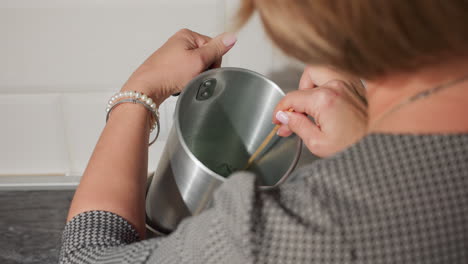 rear view of craft person carefully stirring green liquid substance in metal container placed inside pot on electric stove, using wooden stick attentively during creative preparation