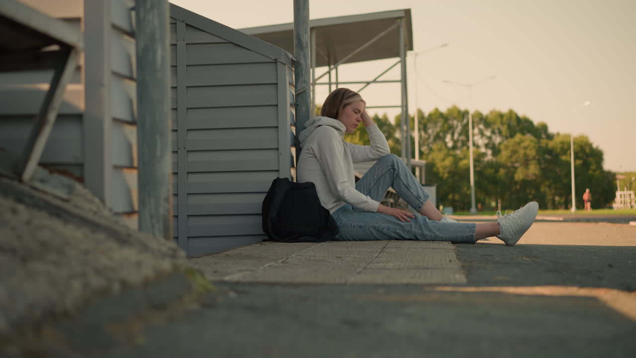 청바지를 입은 소녀가 바닥에 앉아서 머리를 손에 고 검은 가방을 고 깊은 생각에 빠졌고, 배경에서 달리는 누군가의 흐릿한 시야