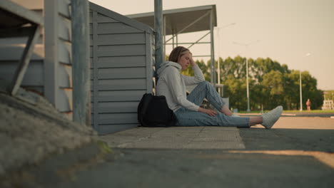 girl in jeans seated on ground, resting head on hand with black bag nearby, deep in thought, blurry view of someone in background running