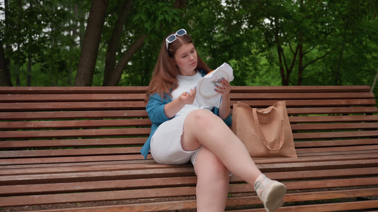 Female stylist sitting on wooden park bench embroidering fabric with needle, wearing blue shirt and white outfit, focused on creative needlework surrounded by peaceful summer greenery
