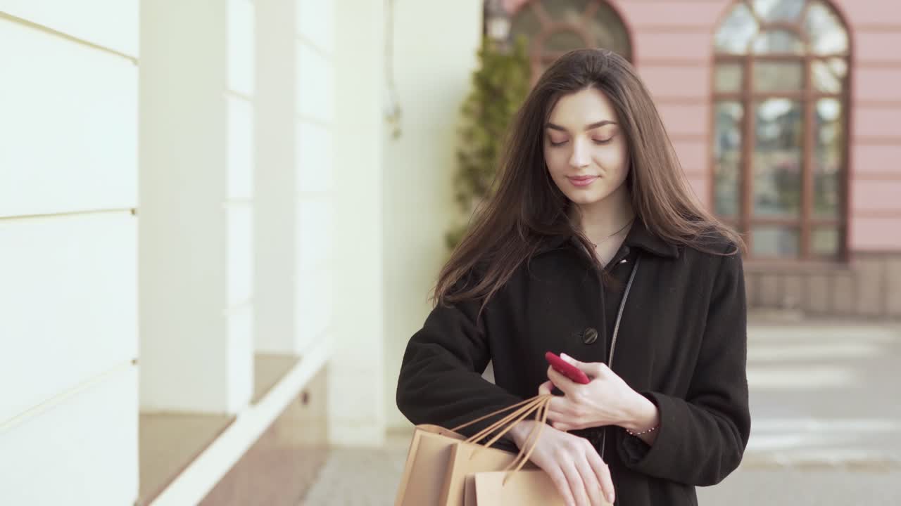 mujer caucásica caminando por la calle con algunas bolsas después de ir de compras girando y mirando la cámara sosteniendo su teléfono inteligente