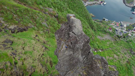 Cinematic-aerial-view-of-a-climber-in-yellow-clothing-at-the-top-of-the-rocky-pinnacle-of-Svolvaergeita-in-spring