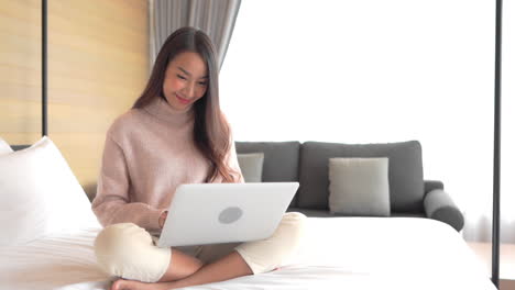 an attractive young woman sits on the bed of her hotel suite as she enters data into her laptop