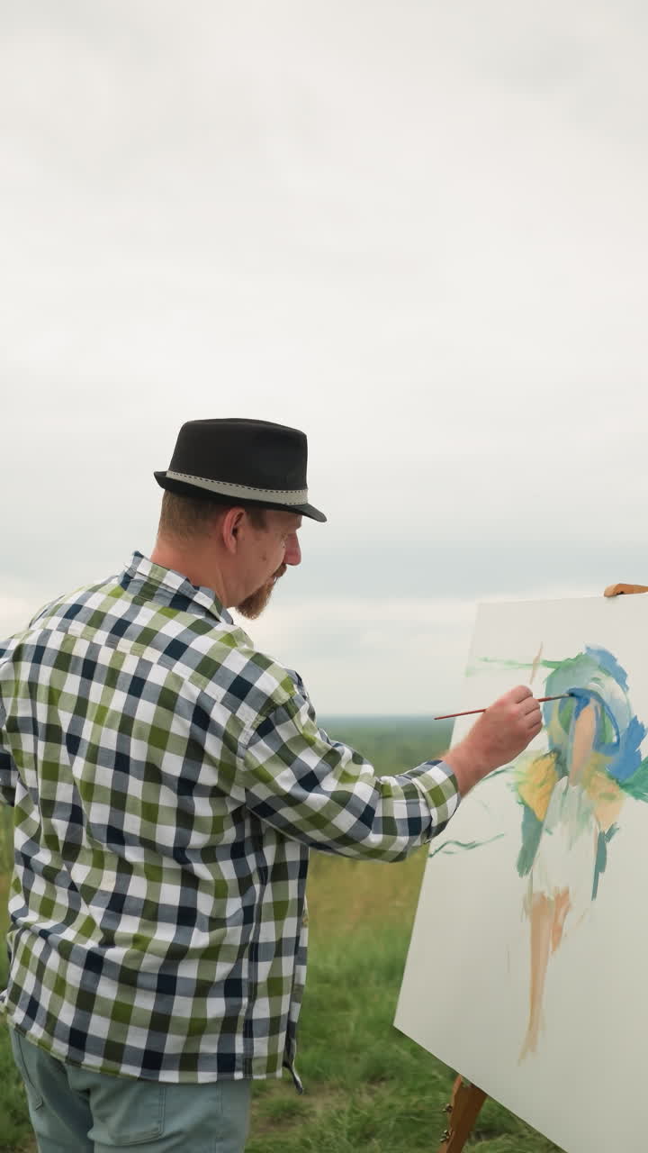 An artist, wearing a hat, checkered shirt, and jeans, is deeply focused on painting on a board in the middle of a lush grassy field under a cloudy sky
