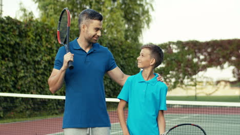 Portrait-Shot-Of-Happy-Father-And-Cute-Teen-Son-Hugging-And-Smiling-Joyfully-At-The-Camera-On-Tennis-Court-In-Summer