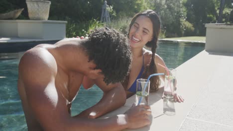 happy biracial couple talking with drinks at pool in garden on sunny day