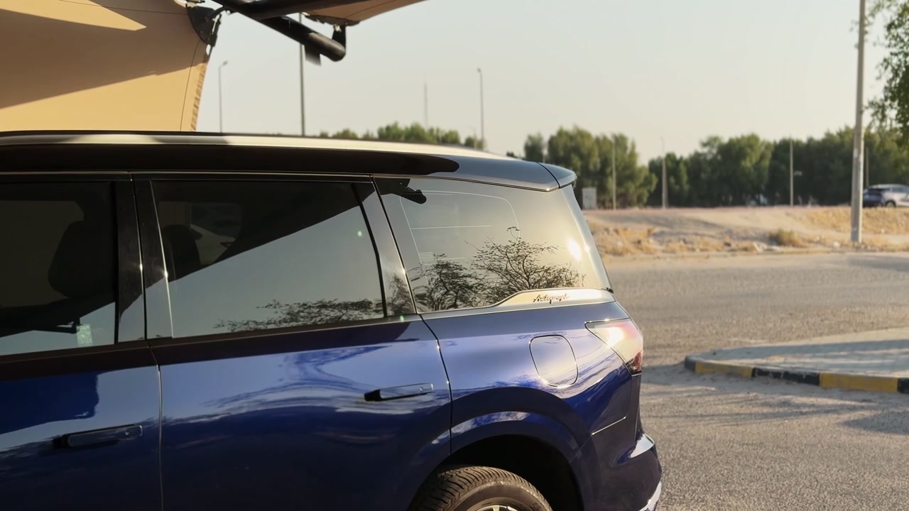 Una vista laterale ravvicinata di un SUV di lusso blu scuro parcheggiato su una strada asfaltata esterna vicino a una banchina sabbiosa. La calda luce dell'ora d'oro si riflette splendidamente sui finestrini oscurati e sulla vernice metallizzata