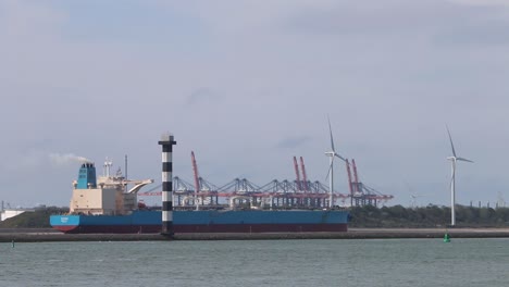 barco comercial en marcha con grúas de astillero y turbinas eólicas en el fondo