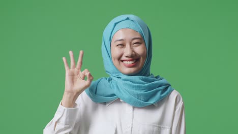 close up of asian muslim woman smiling and showing okay gesture to camera while standing in the green screen background studio