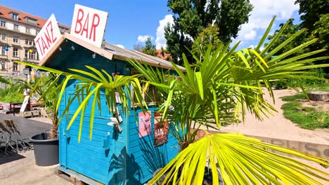 bright green palm leaves move in front of a blue outdoor bar hut on a sandy area, under clear daylight with urban buildings in background