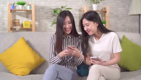 스마트폰을 가진 두 명의 행복한 젊은 아시아 소녀들이 거실의 소파에 앉아 있습니다.