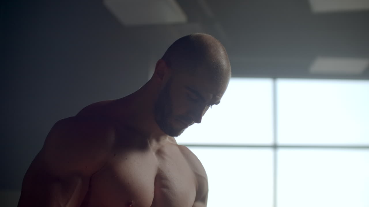 티셔츠를 입지 않은 남자가 기다리고 운동에 적응합니다. 기대. 결정적인 운동 운동 선수를 수행 할 준비가되어 있습니다. 창문 근처에 체육관에 종사하는 강한 남자.