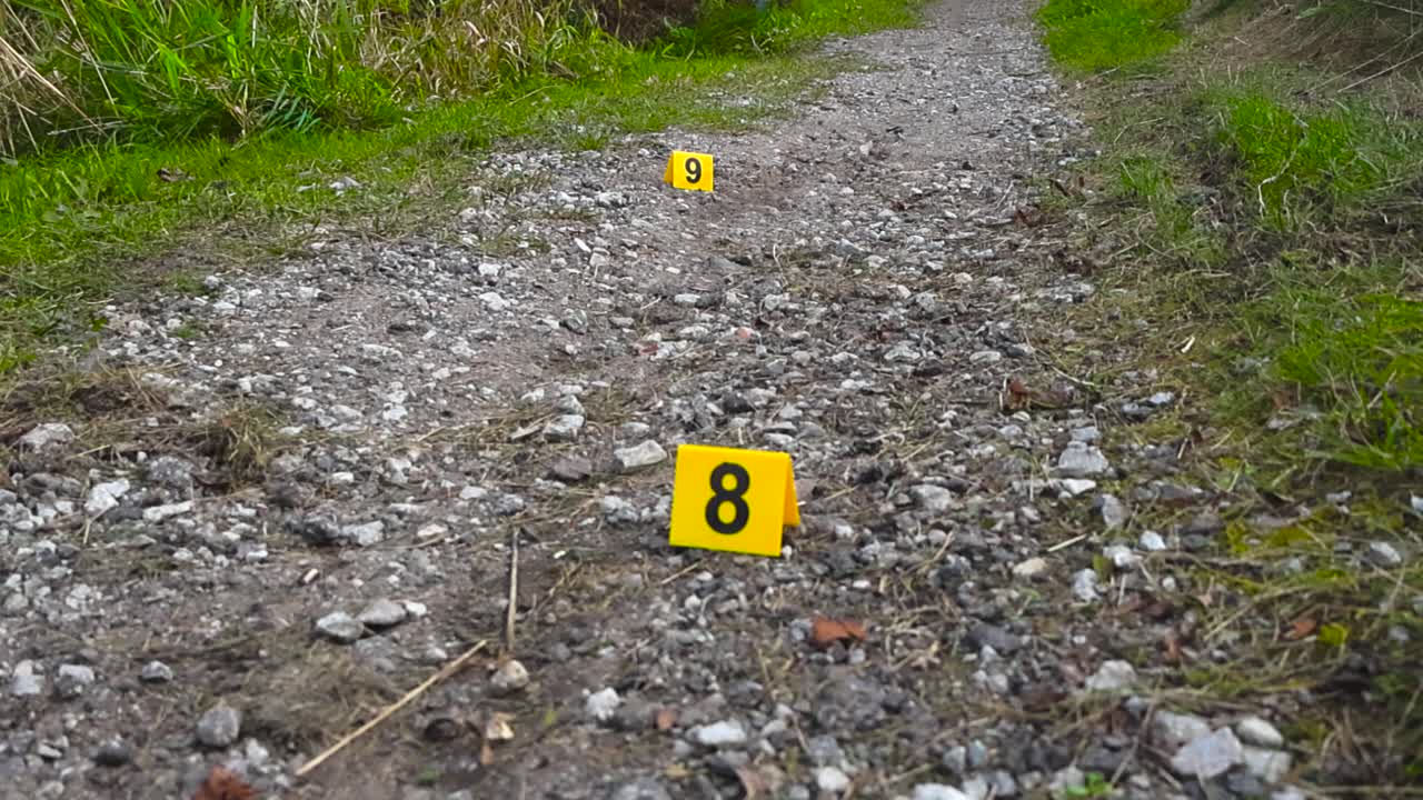 Un movimento di telecamera verso l'alto rivela marcatori numerati accanto a segni di trascinamento su un sentiero di campagna sotto un cielo coperto.