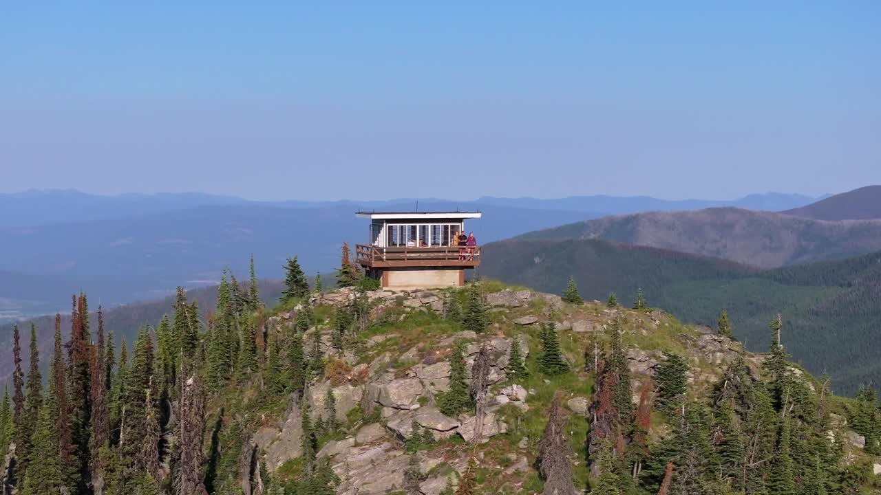 Una ripresa aerea con drone che ruota intorno a una torre di avvistamento antincendio isolata sulla cima di una montagna nel remoto Idaho settentrionale, con un vasto paesaggio aspro sullo sfondo, mentre una coppia si appoggia alla ringhiera