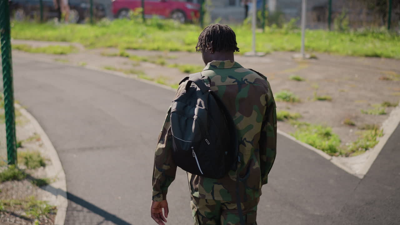 Black Soldier Walking Down Forked Path With Backpack And Camouflage, Back To Camera. Urban Crossroads And Sunlit Pavement Frame Decisive Movement Toward Future And New Direction. Wide Composition