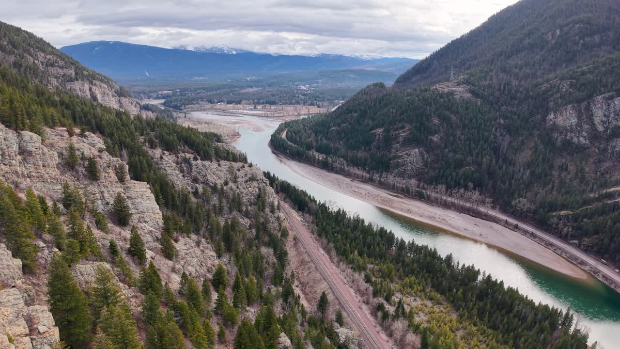 Luftfoto med drone af en turkis flod, der snor sig gennem en dal med klippeskrænter og stedsegrønne skove på den ene side og åbent landbrugsland og bjerge på den anden nær Whitefish, Montana