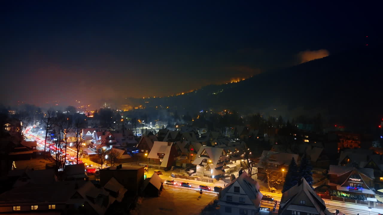 Cozy cottages at the mountain foot lit with lanterns at night. Lots of cars move by the road crossing Zakopane, Poland. Aerial view.