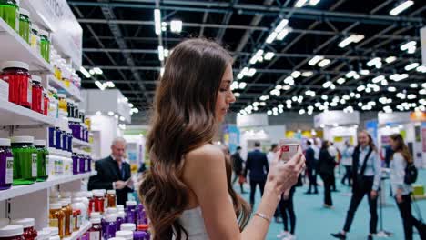 young woman carefully reading labels of various supplements displayed on shelves at a bustling health and wellness expo, demonstrating her interest in healthy lifestyle choices