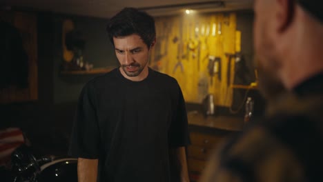 Over-the-shoulder-A-brunette-guy-with-a-shield-hair-in-a-gray-T-shirt-communicates-with-a-mechanic-and-comes-to-an-agreement-in-the-studio-workshop