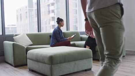 two diverse female work colleagues sitting on sofa holding documents andtablet talking