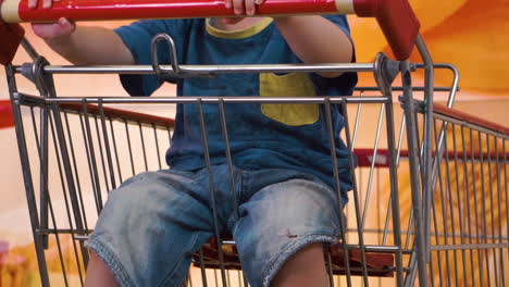 Young-child-in-shopping-cart-inside-store-kicking-feet-camera-pans-up-revealing-boy-waiting-for-parents-alone-and-rocking-cart