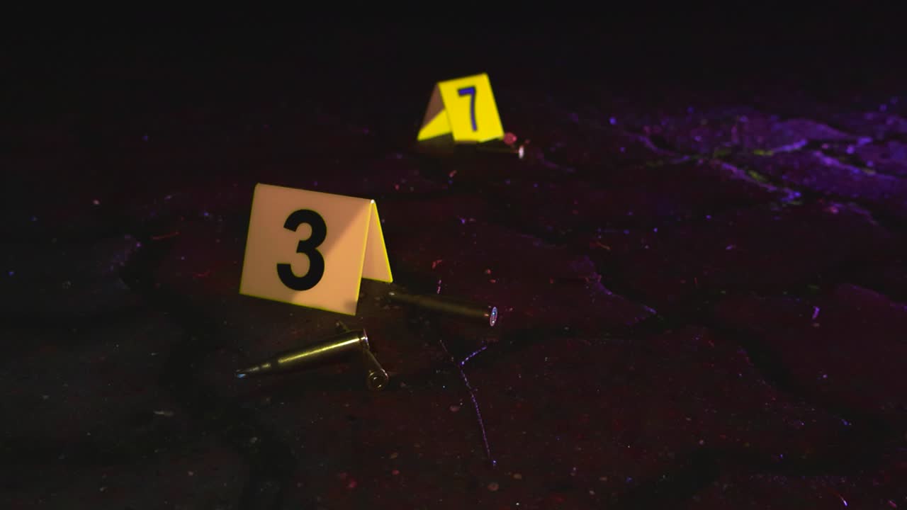 Yellow police numbered crime scene markers next to high caliber cartridges or bullet casings and ammo ammunition on asphalt urban ground while police blue and red lights flash during night time