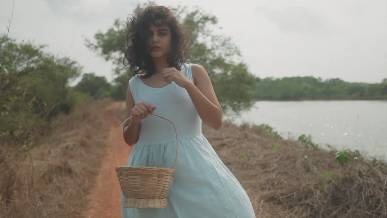 tiro de muñeca de mano en cámara lenta de una modelo india con vestido turquesa y una cesta de madera en la mano parada al lado de un lago en la naturaleza y mirando a la cámara