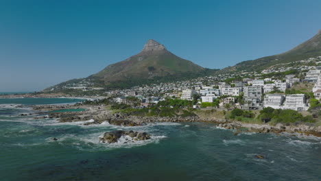 Forwards-fly-above-waves-washing-sea-coast.-Residential-buildings-on-waterfront.-Tall-mountain-with-steep-slopes-and-rock-peak.-Clear-blue-sky.-Cape-Town,-South-Africa