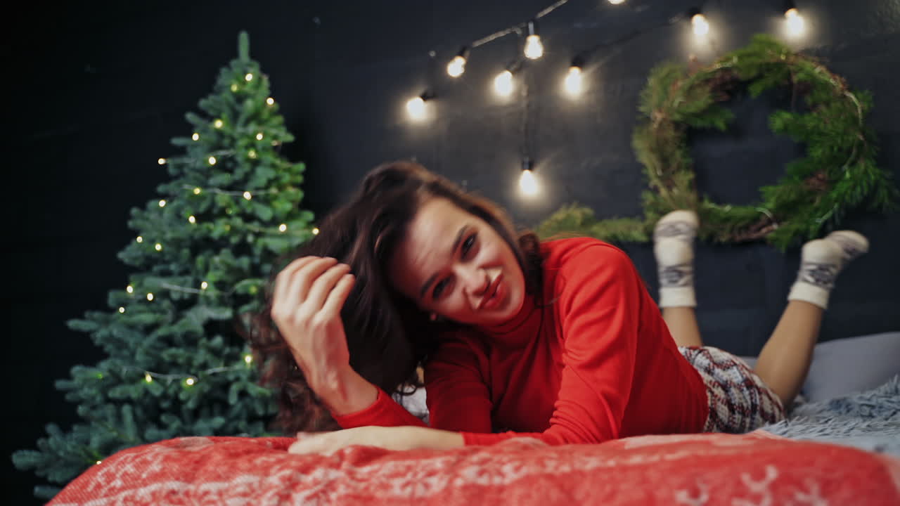 Happy girl before holidays poses on bed and rolling. Beauty in red sweater at Christmas time. Concept video.