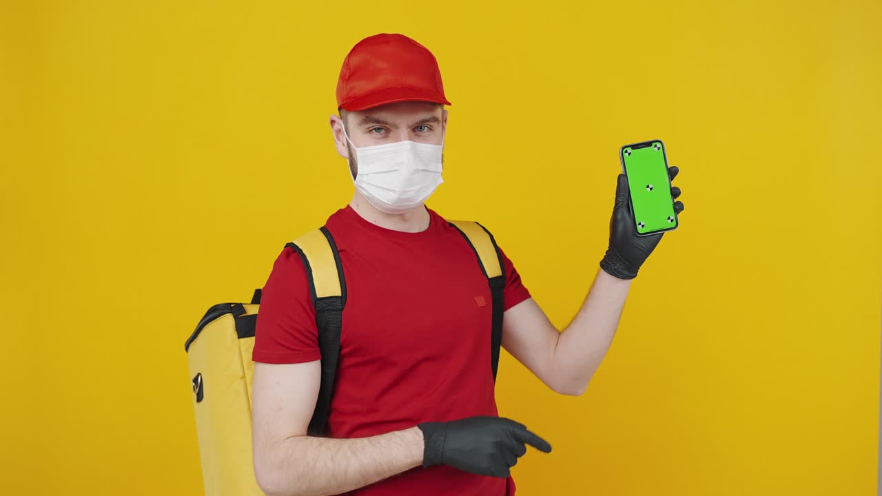 Young delivery man in uniform wearing protective face mask showing cellphone with green chroma key screen for mockup