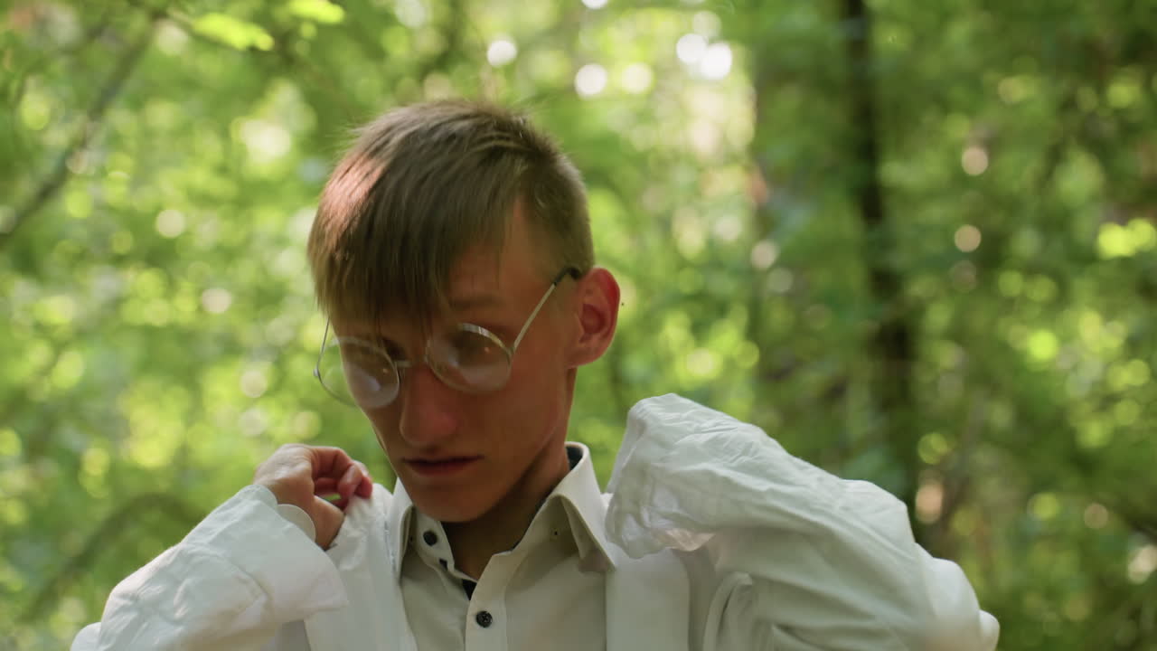 Young biologist in glasses adjusting white coat in forest, showing expression of relief and focus, surrounded by bright green foliage and natural light during outdoor scientific observation