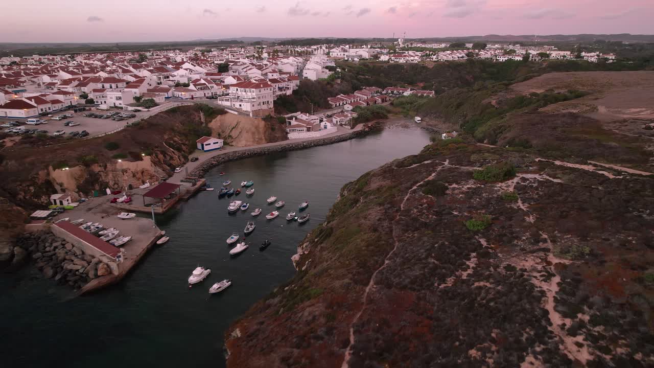Marina de Porto Covo com barcos de pesca ancorados, situada abaixo de uma vila caiada, ao longo das falésias atlânticas escarpadas da Costa Vicentina no Alentejo, Portugal, com aproximação de drone.