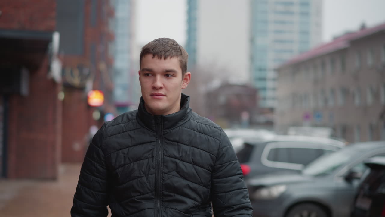 Young automobile enthusiast in black quilted winter coat walking along urban pavement past row of parked vehicles beside brick buildings with blurred city background on overcast gray day