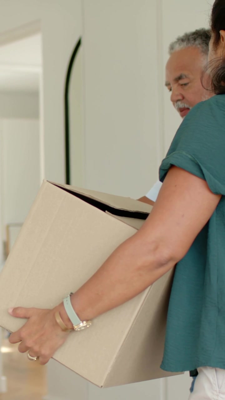 Vertical video: Carrying mature pair angling large box through entryway, woman wearing green blouse