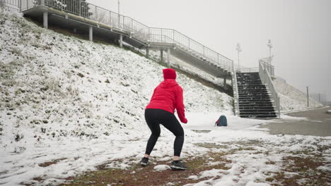 back view of woman performing jump squats during winter outdoor workout on snowy ground, wearing red hoodie and black gloves, scene includes snowy hill with iron rail, foggy atmosphere
