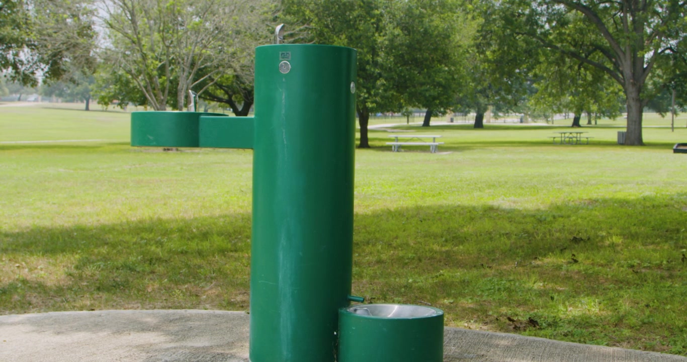 Un'inquadratura ampia di una fontana d'acqua in un parco cittadino vuoto, che evidenzia l'ambiente sereno. La scena cattura la bellezza della fontana sullo sfondo della tranquilla atmosfera del parco.