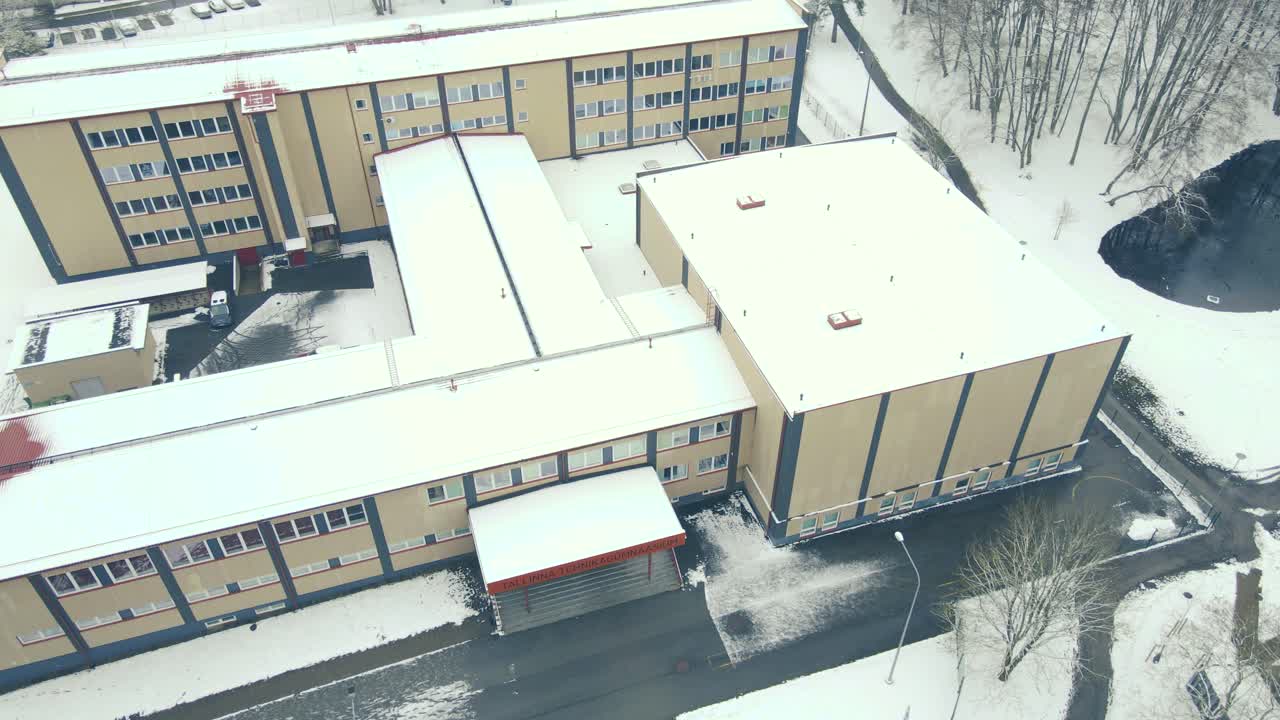Aerial drone footage of a Schoo during winter time named Tallinn Tehnika Gümnaasium or high school during a cloudy dat with white snow around anr on the roof. Tall trees are surrounding in Mustamäe.