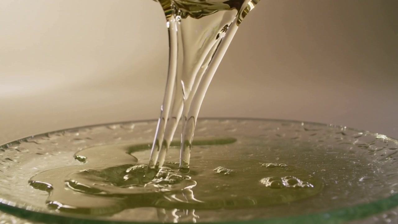Inclinando un beccuccio curvo, un liquido viscoso viene versato in un piatto di vetro poco profondo, formando increspature.