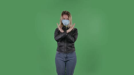 portrait of beautiful young woman in medical protective face mask looking at camera and strictly gesticulates with hands crossed making x shape meaning denial saying no and stop. blonde with loose hair in a black jacket and jeans on a green screen in the