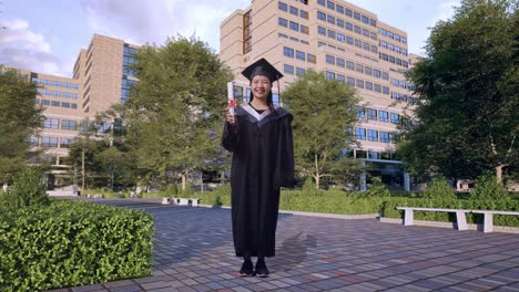 full body of asian woman student graduates in cap and gown smiling and showing diploma in her hand to camera in front of a magnificent university building