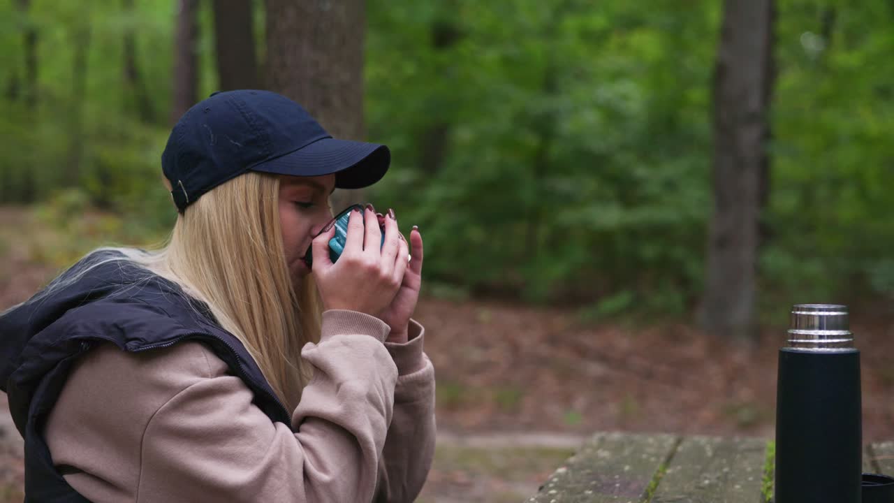 A young woman with long blonde hair drinks from a blue mug while sitting at a wooden table in the forest. Dressed in a cap, sweatshirt, and vest, she enjoys a peaceful moment surrounded by nature