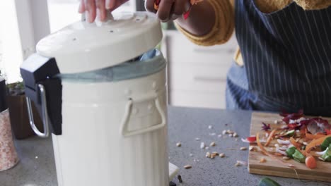 Midsection-of-african-american-woman-in-apron-composting-vegetable-waste-in-kitchen,-slow-motion