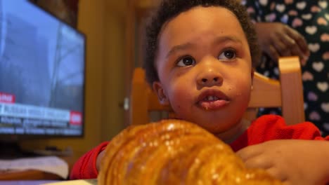 un bebé negro de dos años intenta comer un croissant grande solo en casa sentado al lado de la televisión