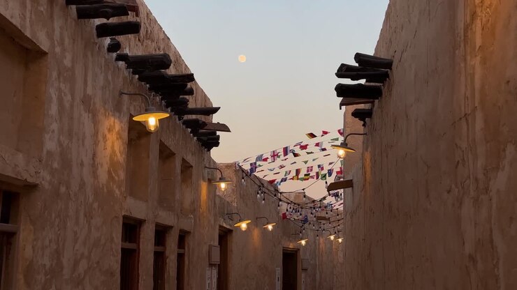 Il vento fa sventolare la bandiera nazionale sopra la stretta via del centro storico di Doha, in Qatar, in un momento dorato al tramonto, tra case in mattoni di fango ristrutturate con strutture in legno e un vicolo acciottolato illuminato di notte.