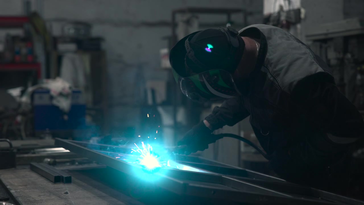 Worker in helmet, heavy jacket, cap, and gloves positions welding torch over steel beams on concrete workbench inside dim industrial workshop, with orange embers, smoky air, and muted metallic